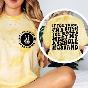 May include: Yellow tie-dye t-shirt with black text and graphics. The front of the shirt has a circular graphic with a skeleton hand making a peace sign and the text "Just a Bitch Married to an Asshole." The back of the shirt says, "If you think I'm a bitch you should meet my asshole husband."