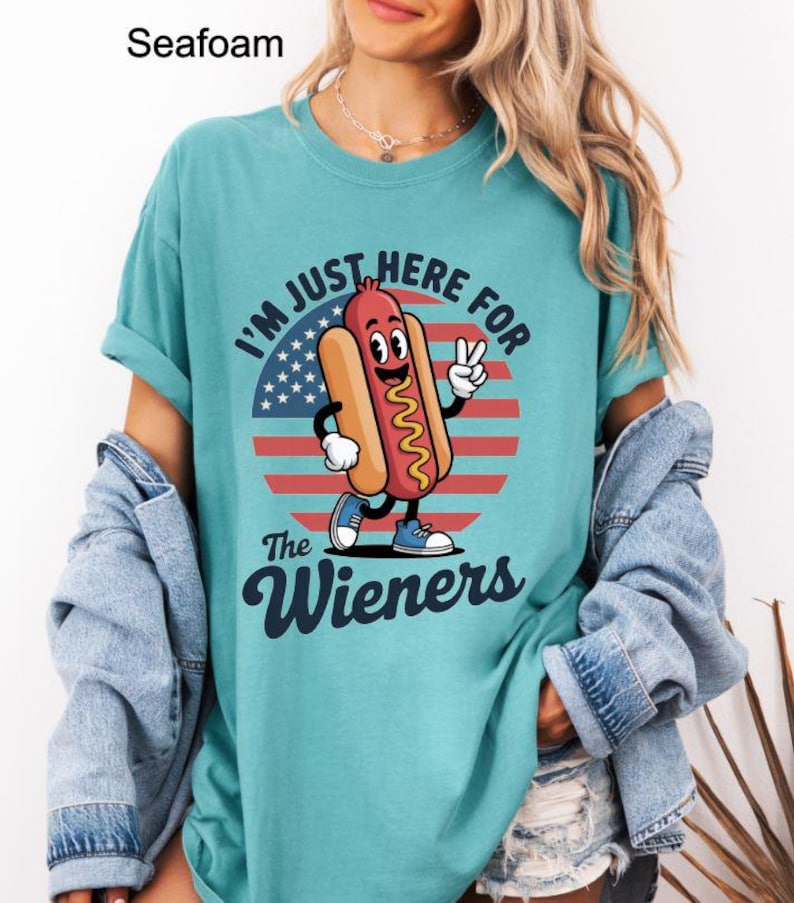 May include: Seafoam green t-shirt with a cartoon hot dog graphic. The hot dog is smiling and giving a peace sign in front of a red, white, and blue design. Text on the shirt reads "I'm Just Here For The Wieners."