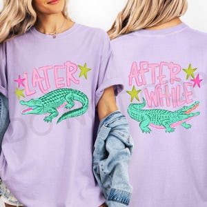 May include: A light purple t-shirt featuring a green alligator graphic with the text "LATER" on the front and "AFTER WHILE" on the back. The design includes pink and yellow stars. The shirt has short sleeves.