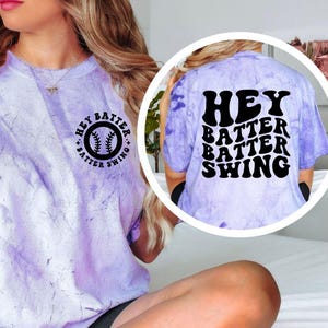 K&ouml;nnte beinhalten: Ein helllila-farbenes T-Shirt mit Batikmuster und dem Text "Hey Batter Batter Swing" auf der Vorder- und R&uuml;ckseite. Die Vorderseite zeigt eine Baseball-Grafik. Auf der R&uuml;ckseite des Shirts steht der Text "Hey Batter Batter Swing" in einem gestapelten Design.