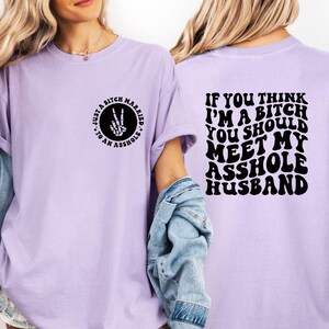 May include: Light purple t-shirt with black text and graphics. The front features a circular design with the text "Just a Bitch Married to an Asshole" and a hand making a peace sign. The back says "If you think I'm a bitch you should meet my asshole husband."