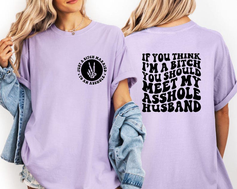 May include: Light purple t-shirt with black text and graphics. The front features a circular design with the text "Just a Bitch Married to an Asshole" and a hand making a peace sign. The back says "If you think I'm a bitch you should meet my asshole husband."