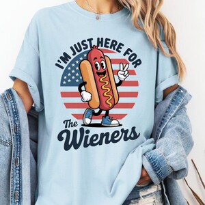 May include: Light blue t-shirt with the text "I'M JUST HERE FOR The Wieners" and a cartoon hot dog character. The hot dog is wearing blue shoes and giving a peace sign. The background features an American flag design.
