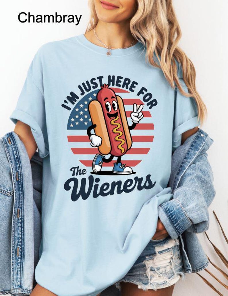 May include: Light blue t-shirt with the text "I'M JUST HERE FOR The Wieners" and a cartoon hot dog character. The hot dog is wearing blue shoes and giving a peace sign. The background features an American flag design.