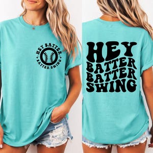 K&ouml;nnte beinhalten: T&uuml;rkisfarbenes T-Shirt mit dem schwarzen Text "HEY BATTER BATTER SWING". Auf der Vorderseite des Shirts befindet sich eine Baseball-Grafik mit demselben Text. Das Shirt hat einen l&auml;ssigen, bequemen Stil.