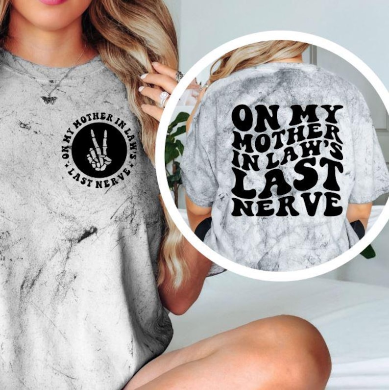 May include: A gray tie-dye t-shirt with the text "ON MY MOTHER IN LAW'S LAST NERVE" in black. The front of the shirt has a black circle with the text "ON MY MOTHER IN LAW'S" and a skeleton hand making a peace sign.