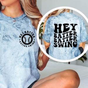 K&ouml;nnte beinhalten: Hellblaues T-Shirt mit Batikmuster und dem Text "Hey Batter Batter Swing" auf der Vorder- und R&uuml;ckseite. Die Vorderseite zeigt eine Baseball-Grafik. Auf der R&uuml;ckseite steht "Hey Batter Batter Swing" in fetter, schwarzer Schrift.