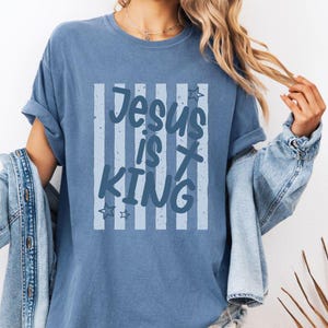 May include: A blue jean-coloured t-shirt featuring the text "Jesus is King" within a striped design. The shirt has rolled-up sleeves and a relaxed fit. The text includes star accents.