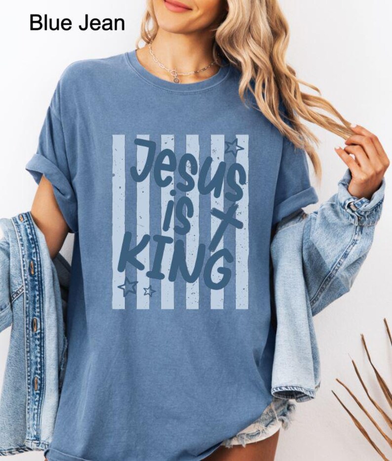 May include: A blue jean-coloured t-shirt featuring the text "Jesus is King" within a striped design. The shirt has rolled-up sleeves and a relaxed fit. The text includes star accents.