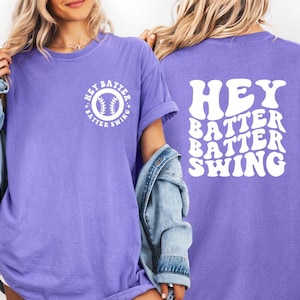 K&ouml;nnte beinhalten: Ein lila T-Shirt mit dem wei&szlig;en Schriftzug "HEY BATTER BATTER SWING" auf dem R&uuml;cken. Auf der Vorderseite befindet sich eine Baseball-Grafik mit dem Text "HEY BATTER SWING" in einem Kreis. L&auml;ssiger Stil.