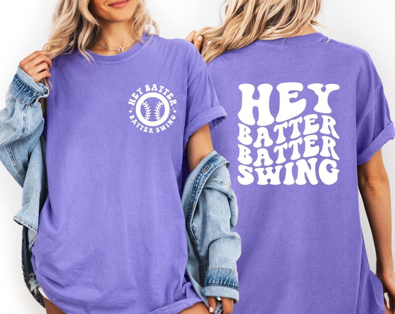 K&ouml;nnte beinhalten: Ein lila T-Shirt mit dem wei&szlig;en Schriftzug "HEY BATTER BATTER SWING" auf dem R&uuml;cken. Auf der Vorderseite befindet sich eine Baseball-Grafik mit dem Text "HEY BATTER SWING" in einem Kreis. L&auml;ssiger Stil.