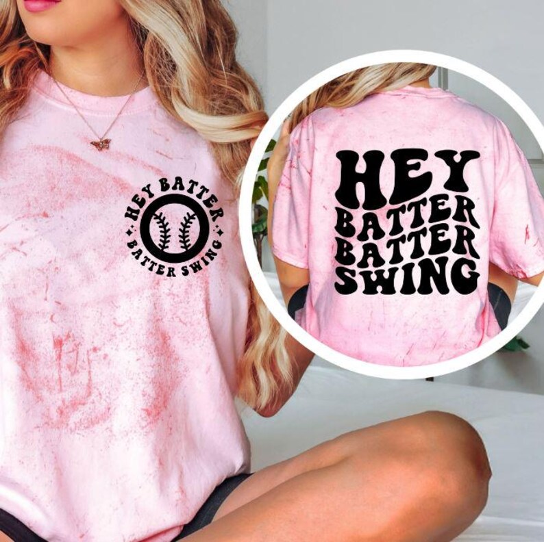 K&ouml;nnte beinhalten: Hellrosa T-Shirt mit Baseball-Design. Auf der Vorderseite des Shirts befindet sich eine runde Grafik mit einem Baseball und den Worten "Hey Batter Swing". Auf der R&uuml;ckseite des Shirts stehen die Worte "Hey Batter Batter Swing".