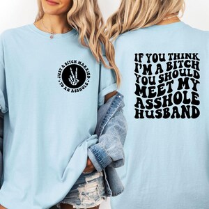 May include: Light blue t-shirt with black text and graphics. The front of the shirt has a circular design with the text "Just a Bitch Married to an Asshole." The back of the shirt reads "If you think I'm a bitch you should meet my asshole husband."