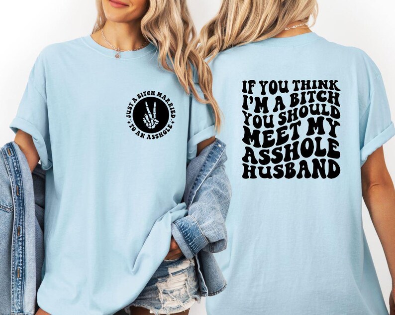 May include: Light blue t-shirt with black text and graphics. The front of the shirt has a circular design with the text "Just a Bitch Married to an Asshole." The back of the shirt reads "If you think I'm a bitch you should meet my asshole husband."