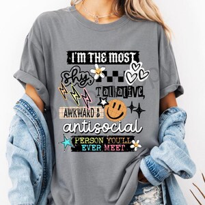 K&ouml;nnte beinhalten: Graues T-Shirt mit dem Text "I'M THE MOST shy, talkative, awkward & antisocial PERSON YOU'LL EVER MEET." Das Design beinhaltet Blitze, Blumen, Herzen und ein Smiley-Gesicht.