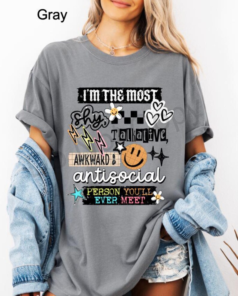 K&ouml;nnte beinhalten: Graues T-Shirt mit dem Text "I'M THE MOST shy, talkative, awkward & antisocial PERSON YOU'LL EVER MEET." Das Design beinhaltet Blitze, Blumen, Herzen und ein Smiley-Gesicht.