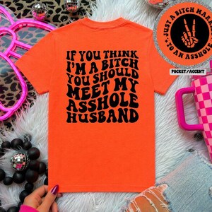 May include: An orange t-shirt with the text "IF YOU THINK I'M A BITCH YOU SHOULD MEET MY ASSHOLE HUSBAND" in black. The shirt is displayed on a white surface with decorative elements.