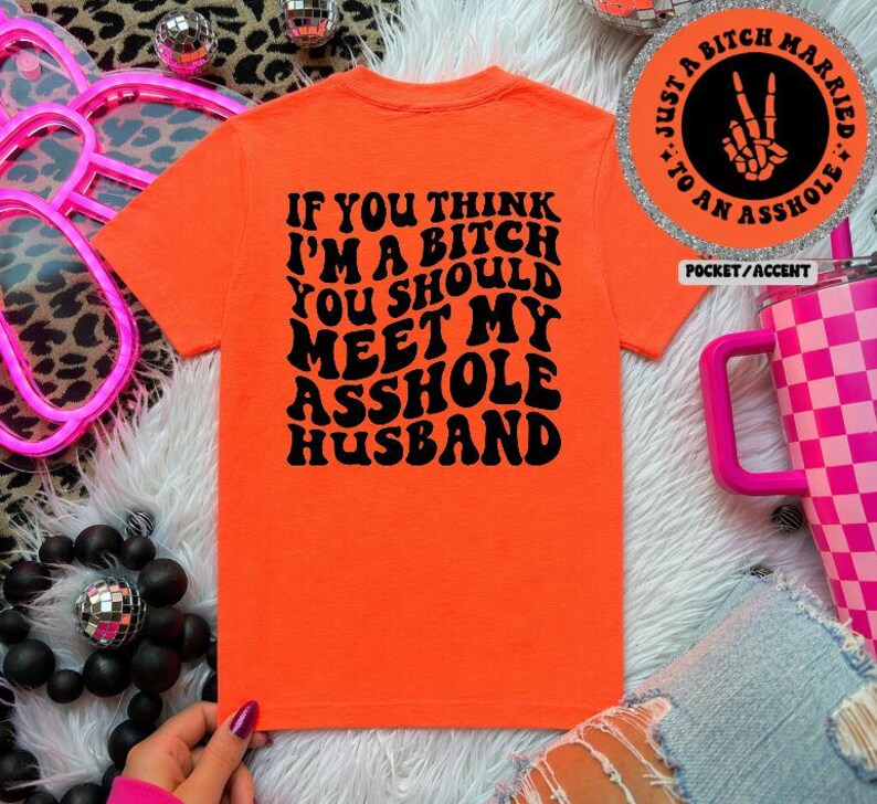 May include: An orange t-shirt with the text "IF YOU THINK I'M A BITCH YOU SHOULD MEET MY ASSHOLE HUSBAND" in black. The shirt is displayed on a white surface with decorative elements.