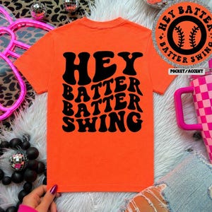 K&ouml;nnte beinhalten: Leuchtend orangefarbenes T-Shirt mit dem Text "HEY BATTER BATTER SWING" in schwarzer Schrift im Retro-Stil. Ein kreisf&ouml;rmiger Aufn&auml;her mit einem Baseball-Motiv und den Worten "HEY BATTER SWING" ist ebenfalls sichtbar. Es ist ein Freizeitkleidungsst&uuml;ck.