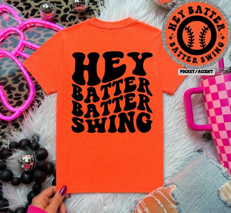 K&ouml;nnte beinhalten: Leuchtend orangefarbenes T-Shirt mit dem Text "HEY BATTER BATTER SWING" in schwarzer Schrift im Retro-Stil. Ein kreisf&ouml;rmiger Aufn&auml;her mit einem Baseball-Motiv und den Worten "HEY BATTER SWING" ist ebenfalls sichtbar. Es ist ein Freizeitkleidungsst&uuml;ck.
