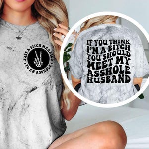 May include: A gray tie-dye t-shirt with black text and graphics. The front of the shirt has a circular design with the words "Just a Bitch Married to an Asshole" and a peace sign. The back of the shirt says "If you think I'm a bitch you should meet my asshole husband."