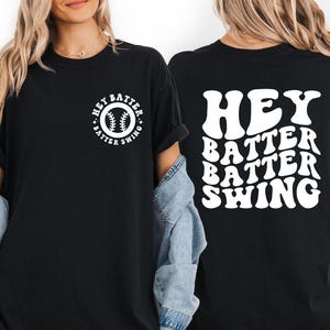 K&ouml;nnte beinhalten: Schwarzes T-Shirt mit wei&szlig;em Text und Baseball-Motiv. Auf der Vorderseite befindet sich ein kreisf&ouml;rmiges Design mit einem Baseball und den Worten "Hey Batter Swing". Auf der R&uuml;ckseite des Shirts steht "Hey Batter Batter Swing" in einer wellenf&ouml;rmigen Schrift.