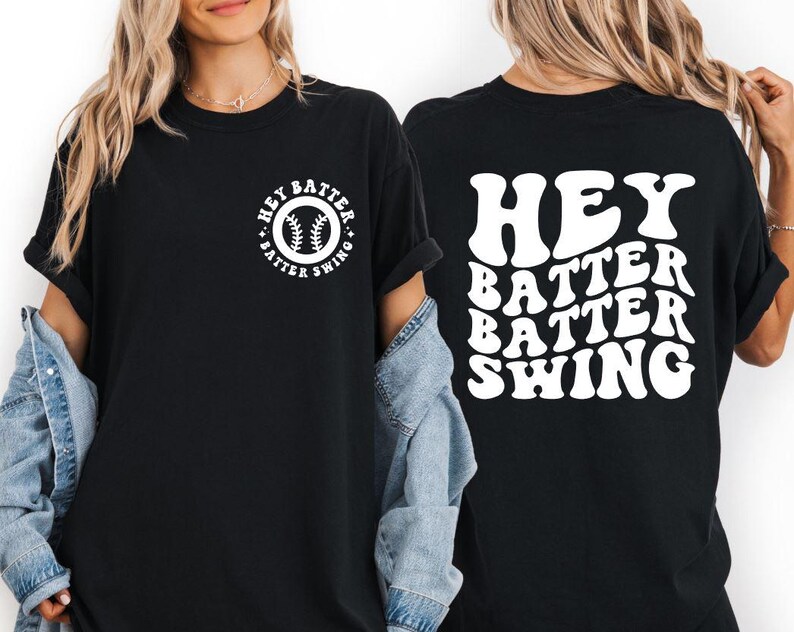 K&ouml;nnte beinhalten: Schwarzes T-Shirt mit wei&szlig;em Text und Baseball-Motiv. Auf der Vorderseite befindet sich ein kreisf&ouml;rmiges Design mit einem Baseball und den Worten "Hey Batter Swing". Auf der R&uuml;ckseite des Shirts steht "Hey Batter Batter Swing" in einer wellenf&ouml;rmigen Schrift.