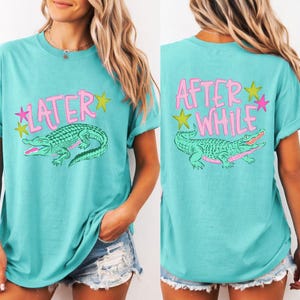 May include: Turquoise t-shirt with a front graphic of a green alligator and the word "LATER" in pink. The back of the shirt features the words "AFTER WHILE" and a similar alligator design. The design includes yellow and pink stars.