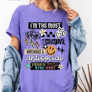 K&ouml;nnte beinhalten: Violettes T-Shirt mit dem Text "I'M THE MOST Shy, Talkative, Awkward, Antisocial PERSON YOU'LL EVER MEET" und verschiedenen Grafiken. Das Shirt hat eine lockere Passform und kurze &Auml;rmel.