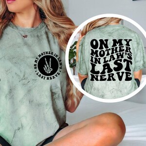 May include: A sage green t-shirt with the text "ON MY MOTHER IN LAW'S LAST NERVE" on the back in black block letters. The front of the shirt has a circular graphic with the text "ON MY MOTHER IN LAW'S LAST NERVE" and a hand making a peace sign.