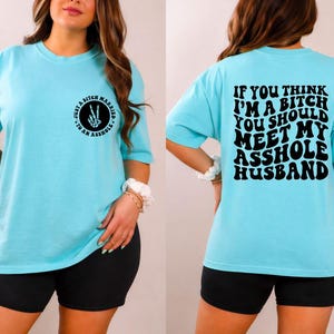 May include: Light blue t-shirt with short sleeves. The front of the shirt has a circular graphic with the text "Just a Bitch Married to an Asshole". The back of the shirt has the text "If you think I'm a bitch you should meet my asshole husband".
