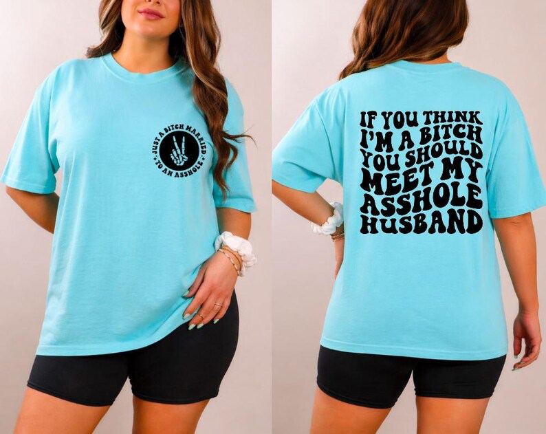 May include: Light blue t-shirt with short sleeves. The front of the shirt has a circular graphic with the text "Just a Bitch Married to an Asshole". The back of the shirt has the text "If you think I'm a bitch you should meet my asshole husband".