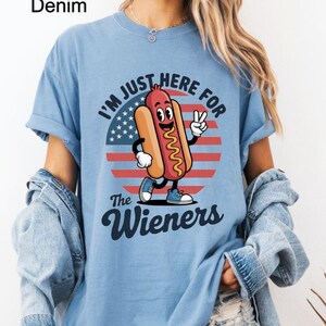 May include: Light blue t-shirt with the text "I'M JUST HERE FOR The Wieners" and a cartoon hot dog graphic. The graphic features a smiling hot dog with a peace sign, set against a red, white, and blue background. The shirt is labeled "Washed Denim".