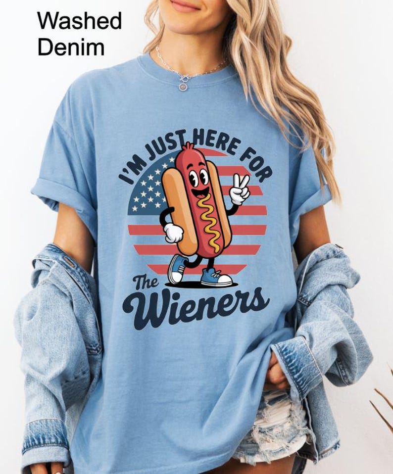 May include: Light blue t-shirt with the text "I'M JUST HERE FOR The Wieners" and a cartoon hot dog graphic. The graphic features a smiling hot dog with a peace sign, set against a red, white, and blue background. The shirt is labeled "Washed Denim".