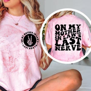 May include: Pink tie-dye t-shirt with the text "ON MY MOTHER IN LAW'S LAST NERVE" in black. The front of the shirt has a circular graphic with the text "ON MY MOTHER IN LAW'S" and a peace sign made of bones.