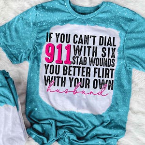 May include: A turquoise t-shirt with white bleach spots and the text "IF YOU CAN'T DIAL 911 WITH SIX STAB WOUNDS YOU BETTER FLIRT WITH YOUR OWN husband" in black, pink, and cursive pink lettering.