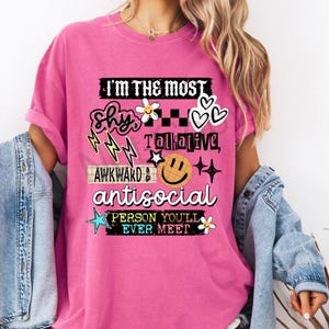 K&ouml;nnte beinhalten: Ein pinkfarbenes T-Shirt mit dem Text "I'M THE MOST shy, awkward, antisocial PERSON YOU'LL EVER MEET." Das Design umfasst Blitze, Blumen, Herzen, Sterne und ein Smiley. Das Shirt ist einfarbig.