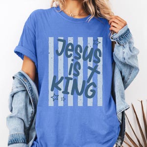 May include: A Flo Blue t-shirt featuring the words "Jesus is King" in a light blue design. The text is set against a background of vertical stripes. The shirt is a solid blue colour.