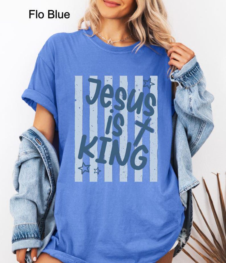 May include: A Flo Blue t-shirt featuring the words "Jesus is King" in a light blue design. The text is set against a background of vertical stripes. The shirt is a solid blue colour.