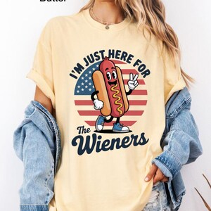 May include: Butter-colored t-shirt with a graphic of a cartoon hot dog character giving a peace sign. The design includes the text "I'M JUST HERE FOR the Wieners" over a red, white, and blue background.