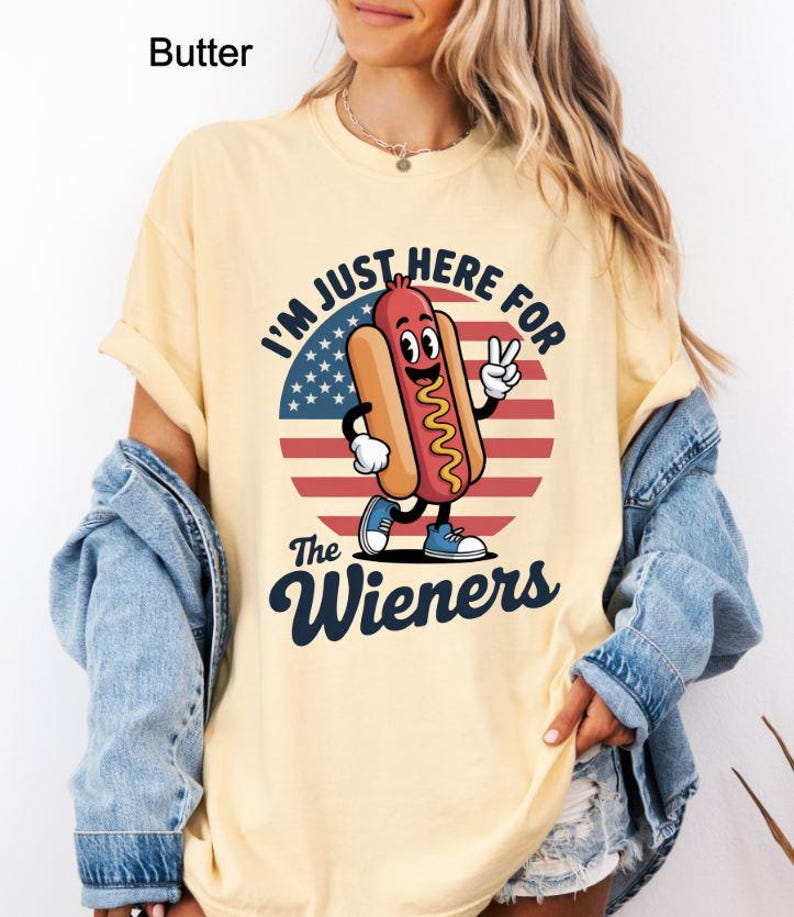 May include: Butter-colored t-shirt with a graphic of a cartoon hot dog character giving a peace sign. The design includes the text "I'M JUST HERE FOR the Wieners" over a red, white, and blue background.