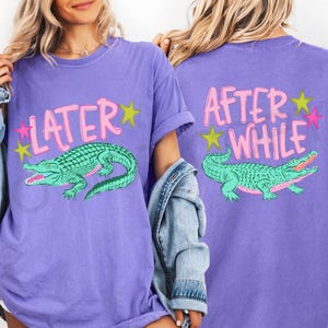May include: Purple t-shirt with a teal alligator graphic and the text "LATER" on the front. The back of the shirt has the text "AFTER WHILE" and a matching alligator graphic. The design includes pink and yellow stars.