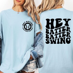 K&ouml;nnte beinhalten: Hellblaues T-Shirt mit dem schwarzen Text "HEY BATTER BATTER SWING". Auf der Vorderseite des T-Shirts befindet sich eine Baseball-Grafik mit dem Text "HEY BATTER SWING". Das T-Shirt hat kurze &Auml;rmel und eine lockere Passform.