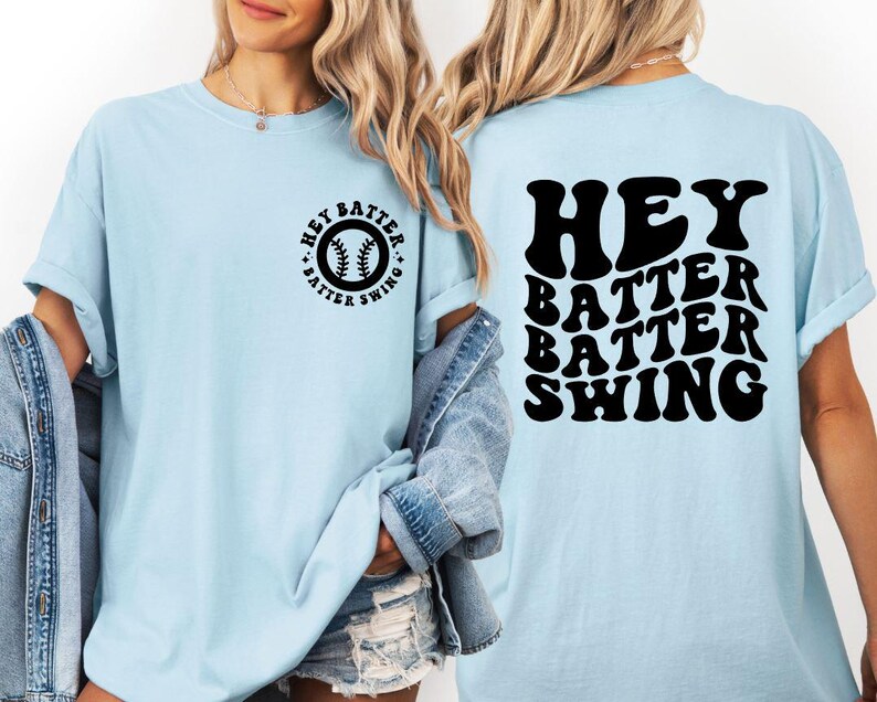 K&ouml;nnte beinhalten: Hellblaues T-Shirt mit dem schwarzen Text "HEY BATTER BATTER SWING". Auf der Vorderseite des T-Shirts befindet sich eine Baseball-Grafik mit dem Text "HEY BATTER SWING". Das T-Shirt hat kurze &Auml;rmel und eine lockere Passform.