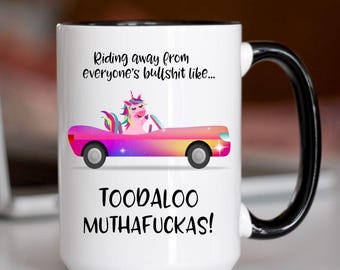 May include: White ceramic mug with a black handle and rim. The mug features the text "Riding away from everyone's bullshit like..." and "TOODALOO MUTHAFUCKAS!". A cartoon unicorn drives a colorful car.