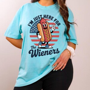 May include: A light blue t-shirt with the text "I'M JUST HERE FOR The Wieners" and a cartoon hot dog character. The hot dog is smiling and giving a peace sign in front of a red, white, and blue graphic.