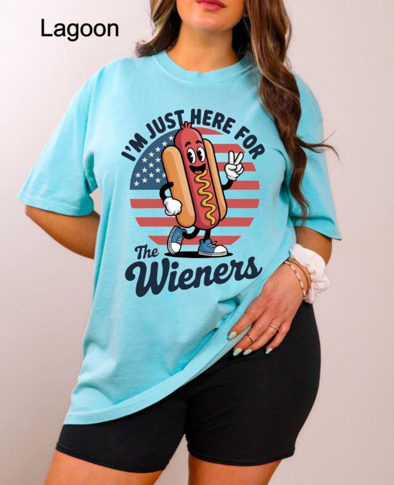 May include: A light blue t-shirt with the text "I'M JUST HERE FOR The Wieners" and a cartoon hot dog character. The hot dog is smiling and giving a peace sign in front of a red, white, and blue graphic.