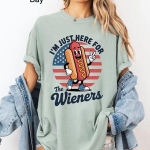 May include: A light green t-shirt featuring a cartoon hot dog character with the text "I'M JUST HERE FOR The Wieners" in a patriotic design. The hot dog is smiling and giving a peace sign. The shirt is a relaxed fit.