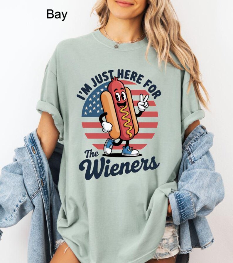 May include: A light green t-shirt featuring a cartoon hot dog character with the text "I'M JUST HERE FOR The Wieners" in a patriotic design. The hot dog is smiling and giving a peace sign. The shirt is a relaxed fit.