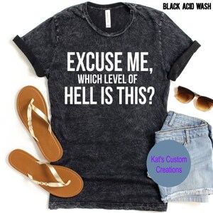 Puede incluir: Camiseta negra con lavado ácido con el texto "EXCUSE ME, WHICH LEVEL OF HELL IS THIS?" Mangas enrolladas, sandalias marrones, gafas de sol y pantalones cortos de mezclilla están colocados alrededor de la camiseta. El texto "Kat's Custom Creations" también es visible.